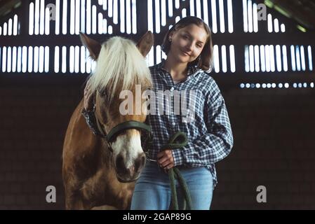 Weibliche Pferdebesitzerin, die mit ihrem hellbraunen Pferd mit einer blonden Mähne im Pferdestall steht. Mädchen hält Bleiseil posiert für die Kamera. Reiten zur freien Verfügung. Stockfoto