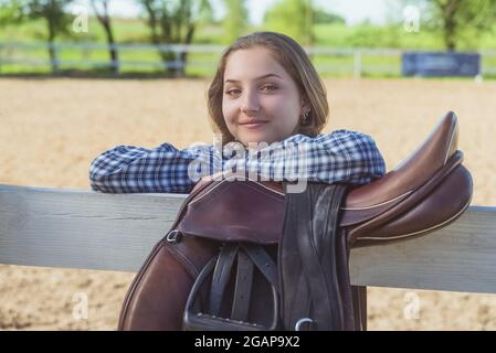 Das junge Mädchen steht mit den Händen auf dem Holzzaun auf der Pferdefarm. Lächelnd und posiert für die Kamera. Ledersattel hängt am Holzzaun im Vordergrund. Stockfoto