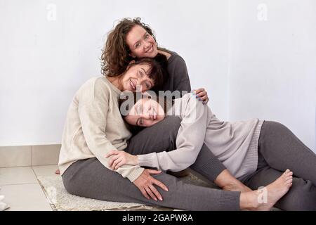 Fröhliche Erwachsene Mutter umarmt charmante junge Töchter, während Pyramide Kopf auf Kopf auf weißem Hintergrund im Studio liegen und auf die Kamera schauen Stockfoto