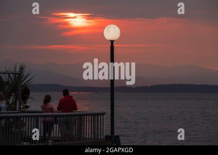 Seattle, USA. 31 Juli 2021. Ein Paar, das einen verrauchten Sonnenuntergang über der Elliott Bay vom Pier 57 aus beobachtet. Klimabrand der Rauch aus den nordwestlichen Waldbränden im pazifik begann gestern auf Seattle zu sinken und erzeugt wunderschöne goldene Sonnenuntergänge. Seattle kämpft jetzt mit rauchigen Himmeln, da jährlich in der Region Klimabrände wüten. James Anderson/ Alamy Live News Stockfoto