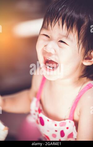 Nahaufnahme bezauberndes asiatisches Kind, das fröhlich lächelt. Nettes chinesisches Mädchen, das Spaß hat und mit Tropfen auf ihrem Gesicht lacht. Tagsüber mit hellem Sonnenlicht. Vintage Stockfoto