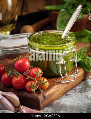 Traditionelle italienische Basilikum-Pesto-Sauce in einem Glas auf einem Holzbrett zusammen mit Basilikum und Tomaten. Stockfoto