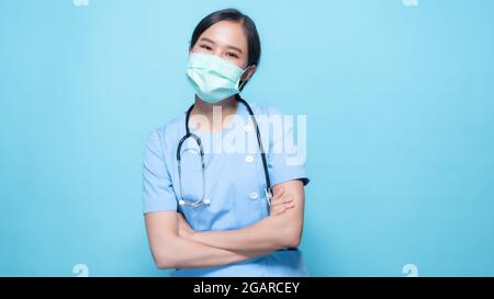 Asiatische Thai Krankenschwester oder Arzt trägt Maske und hält Stethoskop mit großen lächelte isoliert im Studio auf blauem Hintergrund Stockfoto