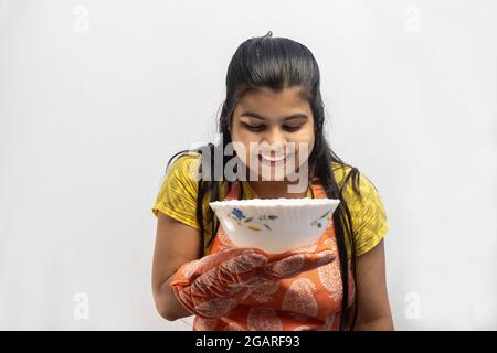 Eine hübsche indische Hausfrau, die Kochschürze und Ofenhandschuhe mit einer Schüssel in der Hand trägt, lächelt auf weißem Hintergrund Stockfoto