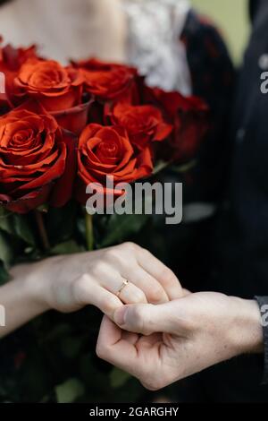 Brautpaar und Bräutigam halten die Hände auf dem Hintergrund eines Blumenstraußes aus roten Rosen, Nahaufnahme von Zärtlichkeit und Romantik. Stockfoto