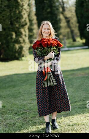 Junges Mädchen, das lächelnd auf dem Rasen steht und mit einem Strauß roter Rosen auf die Kamera blickt Stockfoto