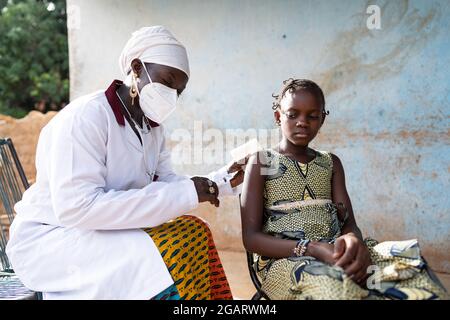 Auf diesem Bild spritzt eine sanfte schwarze Krankenschwester mit einer schützenden Gesichtsmaske zart einen Impfstoff in den Arm eines entspannten kleinen afrikanischen Schulmädchen du Stockfoto