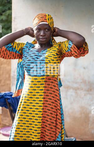 In diesem Bild versucht eine nachdenkliche junge schwarz-afrikanische Schönheit mit erhobenen Armen, ihre Kopfbedeckung anzupassen, die zu ihrem kolossal traditionellen Kleid passt Stockfoto
