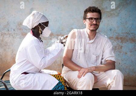 Auf diesem Bild impft eine schwarze Krankenschwester mit einer Gesichtsmaske einen jungen kaukasischen Mann mit einem selbstbewussten Lächeln auf seinem Gesicht gegen Gelbfieber in einem Stockfoto