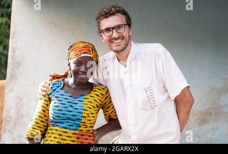 Auf diesem Bild lächelt ein junger weißer Freiwilliger-Arzt fröhlich in die Kamera, während eine wiedergeborene afrikanische Frau in einem traditionellen Kleid und Kopfbedeckung steht Stockfoto