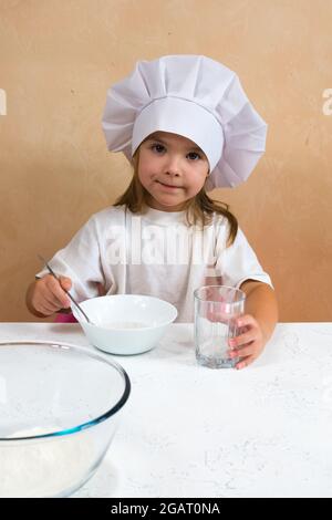 Ein kleines Mädchen, das als Köchin gekleidet ist, knetet den Teig. Konzept zur Entwicklung von Kindern. Die Entwicklung der Feinmotorik der Hände. Das Kind liebt, hat Spaß Stockfoto