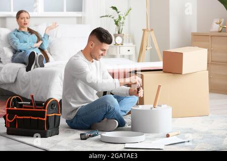 Junger Mann, der Möbel zusammenstellt, während seine faule Frau auf dem Bett liegt Stockfoto