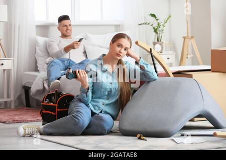 Junge Frau, die Möbel zusammenstellt, während ihr fauler Mann auf dem Bett liegt Stockfoto