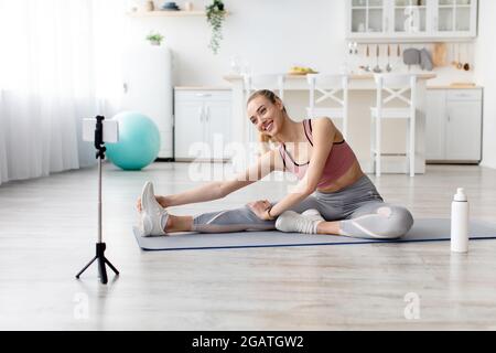 Ferngesteuert trainieren, Fitness-Blogger arbeitet zu Hause, covid-19 Lockdown. Glückliche junge Blondine in Sportswear auf Matte tut Stretching für Beine, schaut am Telefon wir Stockfoto