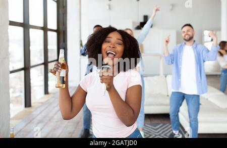 Millennial Black Lady mit Bierflasche und Mikro singen Karaoke für ihre vielfältigen Freunde zu Hause. Schöne afroamerikanische Frau, die Lied aufführt, gibt Stockfoto