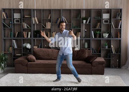 Junger Mann mit Kopfhörern tanzt im Wohnzimmer mit dem Smartphone