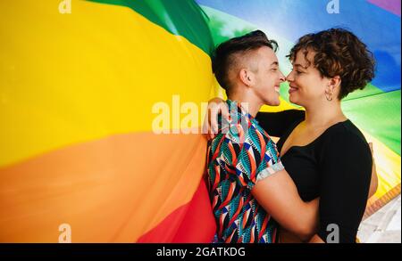 Liebevolles queeres Paar, das sich gegen eine Regenbogenrüde-Flagge festmacht. Das junge LGBTQ-Paar umarmt sich gegenseitig und berührt ihre Nasen. Zwei nicht-con Stockfoto