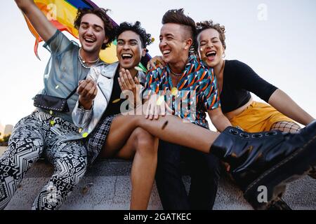 Junge Menschen feiern Stolz, während sie zusammen sitzen. Vier Mitglieder der LGBTQ+ Community lächeln fröhlich und heben die Pride-Flagge. Gruppe von Stockfoto