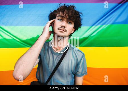 Homosexuell Mann umarmt seine Identität. Junge Schwule, die die Kamera anschaut, während sie gegen eine stolze Flagge steht. Junger queerer Mann mit Lidschatten gegen den LGB Stockfoto
