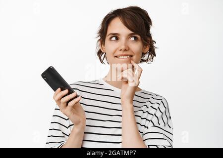 Porträt einer jungen, nachdenklichen Frau, die Mobiltelefon hält, fasziniert lächelt, versucht, smth zu versuchen, eine Idee zu haben, vor weißem Hintergrund stehend Stockfoto