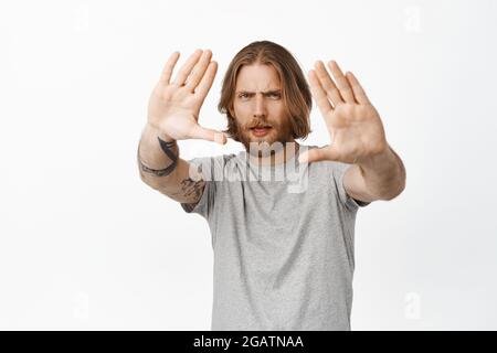 Leidenschaftlicher und kreativer blonder bärtiger Mann mit langen Haaren, der durch Handrahmen schaut und nachdenklich zuckt, interessante Idee schafft, stehend Stockfoto