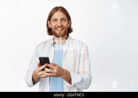 Junger Mann, der Musik über Kopfhörer hört, lächelt, Mobiltelefon hält, Kopfhörer benutzt und vor weißem Hintergrund steht Stockfoto