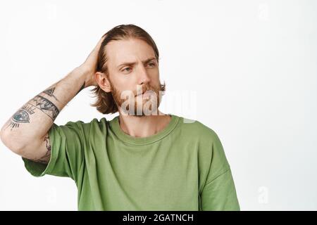 Bild eines frechen, hübschen bärtigen Kerls mit Tattoos, Hipster, der seinen langen Haarschnitt berührt, Finger durch die Haare läuft und zur Seite schaut, gegen den er steht Stockfoto