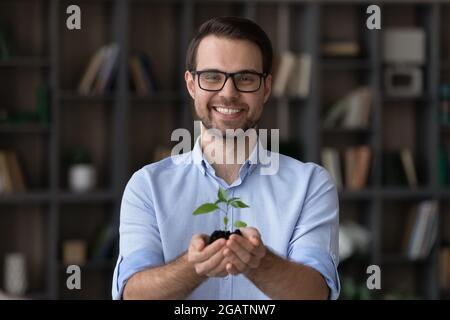 Glücklicher junger Kerl, der lächelnd auf die Kamera schaut und einen kleinen Sämling hält Stockfoto