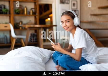 Das Teenager-Mädchen sitzt mit einem Mobiltelefon auf dem Bett und trägt Kopfhörer. Sie hört Musik über eine mobile Online-Anwendung. Das Konzept von Stockfoto