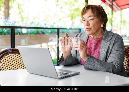 Das Foto einer ernsthaften 60-jährigen Frau arbeitet online im Sommercafé. Sie sitzt vor dem Laptop-Monitor und spricht während eines Online-Videoanrufs. M Stockfoto