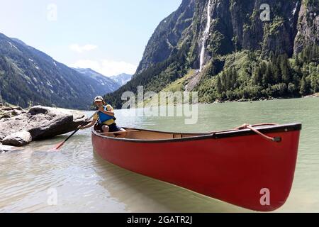 Junge sitzt im Kanu Reiten auf Bergsee in Sommer alpine Landschaft mit Wasserfall in Bacgrund. Stillup Stausee, Stillup Lake, Österreich, Tirol Stockfoto