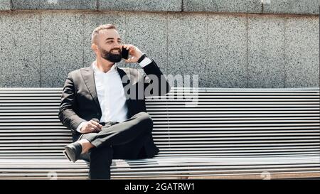 Selbstbewusster, reifer Geschäftsmann, der im Freien mit dem Smartphone spricht, mit dem Kunden auf dem Mobiltelefon in der Nähe des Geschäftszentrums spricht, auf der Bank sitzt i Stockfoto