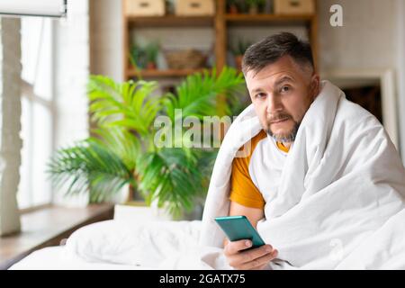 Ein kranker Mann mittleren Alters sitzt auf einem Bett unter einer Decke. Er hält ein Smartphone in der Hand und wird seinen Arzt für eine Online-Konsultation anrufen. Quarantäne Stockfoto