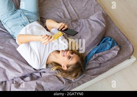 Eine Frau, die im Bett liegt, bestellt über eine mobile Anwendung. Sie wählt die Kartennummer für Online-Einkäufe. Online-Shopping-Konzept. Stockfoto