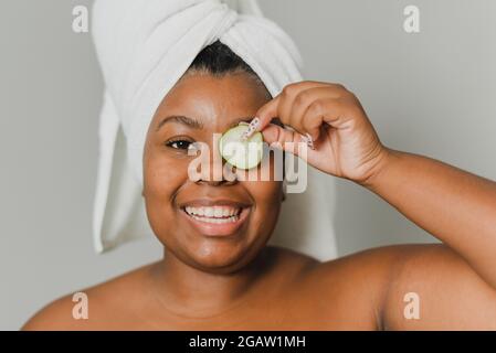 Porträt einer glücklichen jungen schwarzen Frau mit einem Handtuch, das sich mit einer natürlichen Gurke um ihre Haut kümmert. Stockfoto