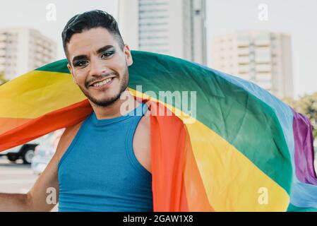 Junger schwuler hispanischer Mann, der auf der Straße eine bunte Flagge trägt, fühlt sich glücklich Stockfoto