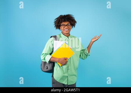 Aufgeregter afroamerikanischer Student, der leeren Raum und lächelnd zeigt, mit Büchern, Notizbüchern und Rucksack posiert, auf blauem Hintergrund stehend, Kopie s Stockfoto