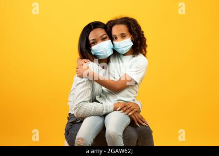 Porträt einer afroamerikanischen Mutter und Tochter mit schützenden medizinischen Masken im Gesicht und Blick auf die Kamera auf gelbem Hintergrund. Schwarze Mama und Stockfoto