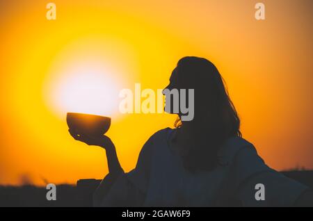 Filmische Silhouette einer Frau, die bei einem wunderschönen orangefarbenen Sonnenuntergang mit Sonnenscheibe eine tibetische Schale spielt. Konzept: Frieden, Ruhe, Zen, Klangheilung Stockfoto