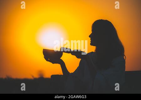 Filmische Silhouette einer Frau, die bei einem wunderschönen orangefarbenen Sonnenuntergang mit Sonnenscheibe eine tibetische Schale spielt. Konzept: Frieden, Ruhe, Zen, Klangheilung Stockfoto