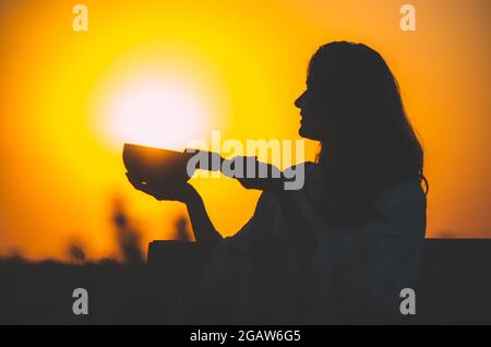 Filmische Silhouette einer Frau, die bei einem wunderschönen orangefarbenen Sonnenuntergang mit Sonnenscheibe eine tibetische Schale spielt. Konzept: Frieden, Ruhe, Zen, Klangheilung Stockfoto