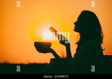 Filmische Silhouette einer Frau, die bei einem wunderschönen orangefarbenen Sonnenuntergang mit Sonnenscheibe eine tibetische Schale spielt. Konzept: Frieden, Ruhe, Zen, Klangheilung Stockfoto