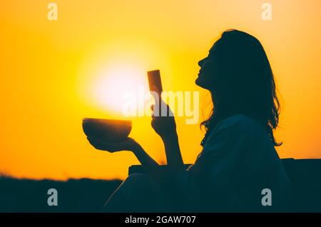 Filmische Silhouette einer Frau, die bei einem wunderschönen orangefarbenen Sonnenuntergang mit Sonnenscheibe eine tibetische Schale spielt. Konzept: Frieden, Ruhe, Zen, Klangheilung Stockfoto