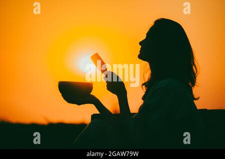 Filmische Silhouette einer Frau, die bei einem wunderschönen orangefarbenen Sonnenuntergang mit Sonnenscheibe eine tibetische Schale spielt. Konzept: Frieden, Ruhe, Zen, Klangheilung Stockfoto