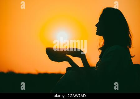 Filmische Silhouette einer Frau, die bei einem wunderschönen orangefarbenen Sonnenuntergang mit Sonnenscheibe eine tibetische Schale spielt. Konzept: Frieden, Ruhe, Zen, Klangheilung Stockfoto