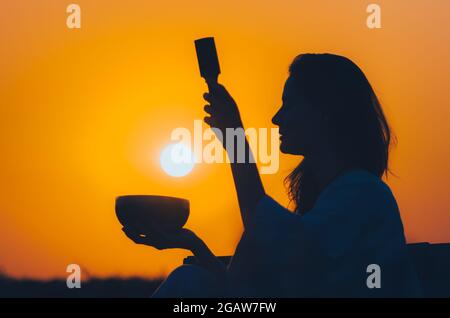 Filmische Silhouette einer Frau, die bei einem wunderschönen orangefarbenen Sonnenuntergang mit Sonnenscheibe eine tibetische Schale spielt. Konzept: Frieden, Ruhe, Zen, Klangheilung Stockfoto