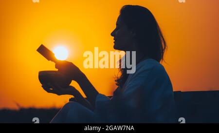 Filmische Silhouette einer Frau, die bei einem wunderschönen orangefarbenen Sonnenuntergang mit Sonnenscheibe eine tibetische Schale spielt. Konzept: Frieden, Ruhe, Zen, Klangheilung Stockfoto