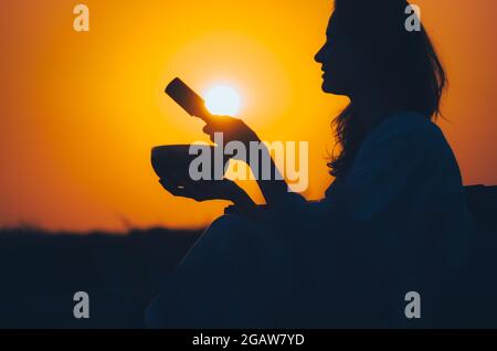 Filmische Silhouette einer Frau, die bei einem wunderschönen orangefarbenen Sonnenuntergang mit Sonnenscheibe eine tibetische Schale spielt. Konzept: Frieden, Ruhe, Zen, Klangheilung Stockfoto