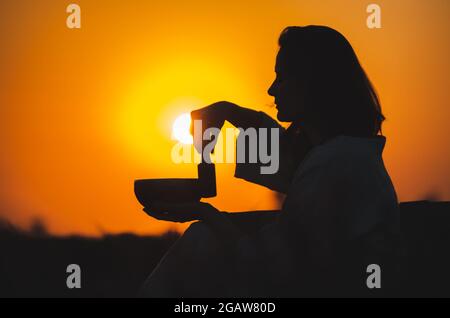 Filmische Silhouette einer Frau, die bei einem wunderschönen orangefarbenen Sonnenuntergang mit Sonnenscheibe eine tibetische Schale spielt. Konzept: Frieden, Ruhe, Zen, Klangheilung Stockfoto