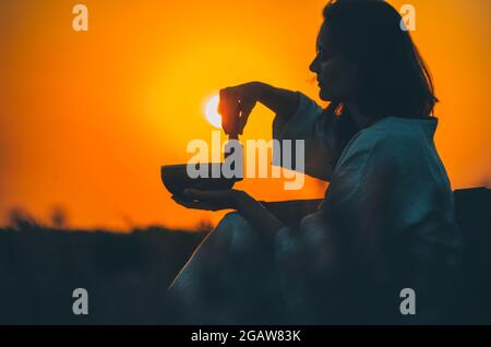 Filmische Silhouette einer Frau, die bei einem wunderschönen orangefarbenen Sonnenuntergang mit Sonnenscheibe eine tibetische Schale spielt. Konzept: Frieden, Ruhe, Zen, Klangheilung Stockfoto